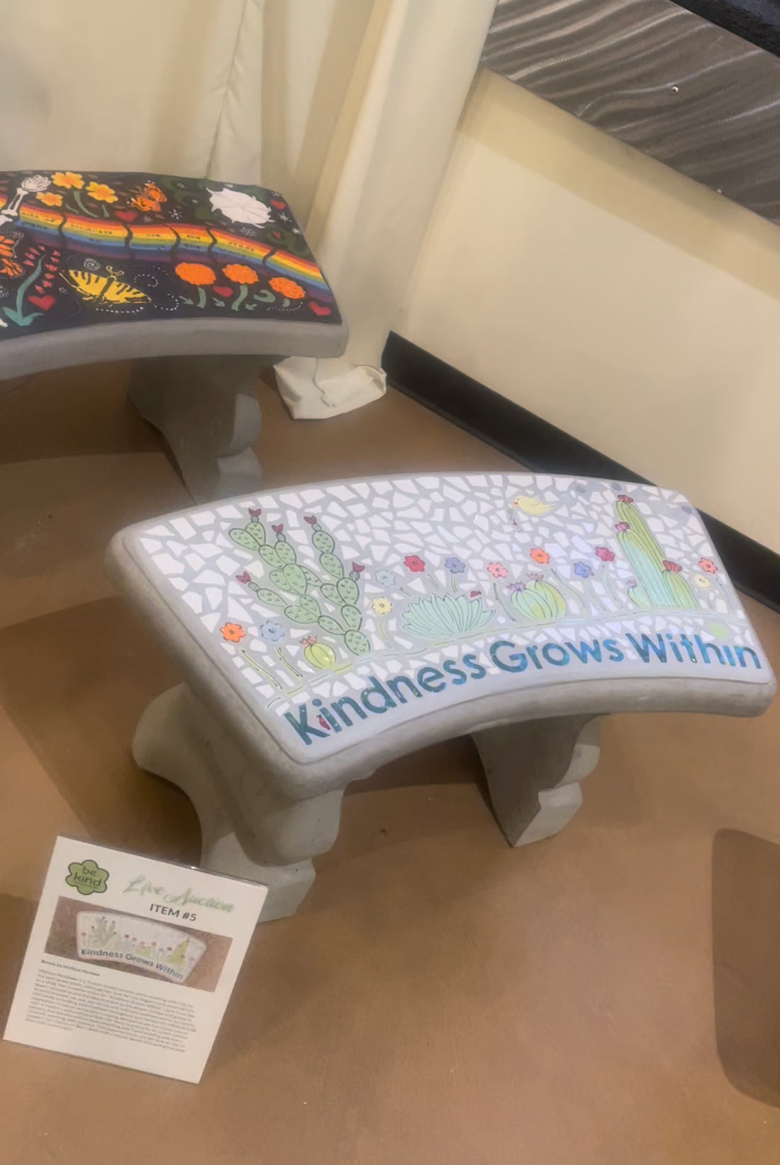 A mosaic bench with cactus, flowers and a white background, with the saying “Kindness Grows Within” across the bottom